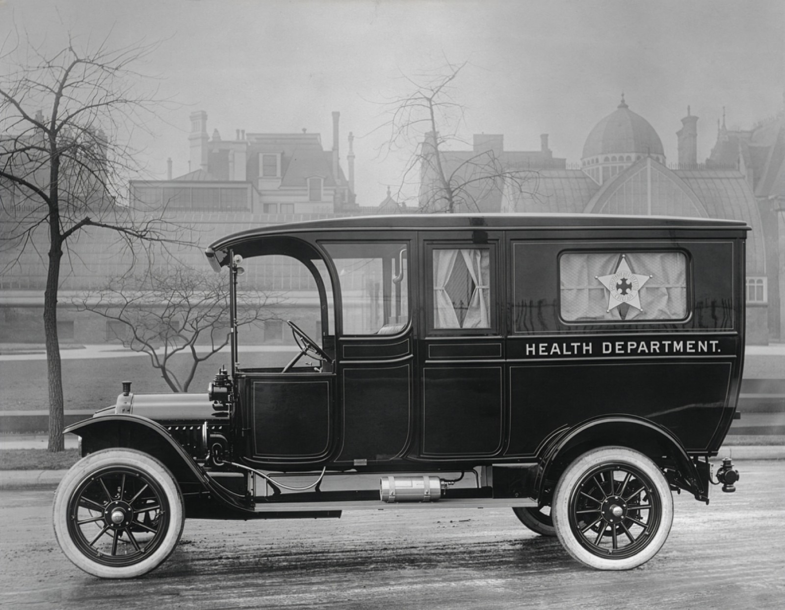 Early 1900s motorized Health Department ambulance with a medical emblem in its rear window, parked on a city street.