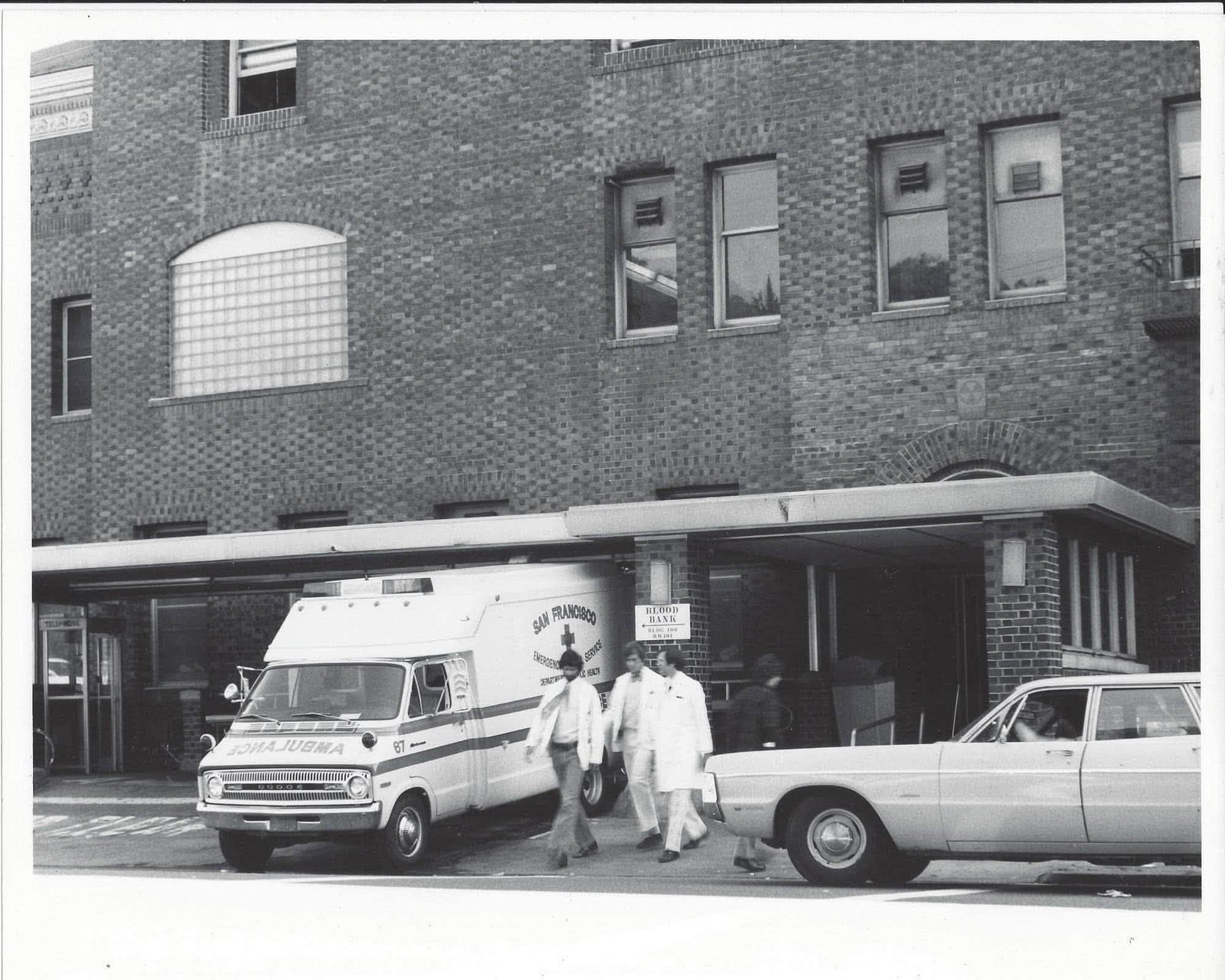 A 1970s Dodge box ambulance lettered San Francisco Emergency Ambulance Service, Department of Public Health, parked at a hospital entrance as staff in white coats walk past